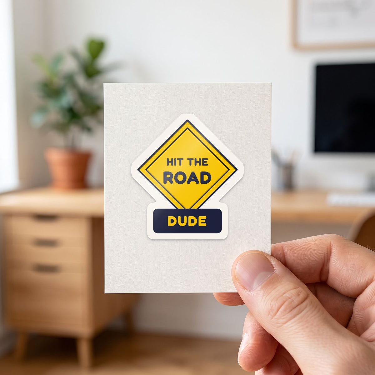 Hit the Road, Dude (Road Sign) sticker held in a hand on its white backing card